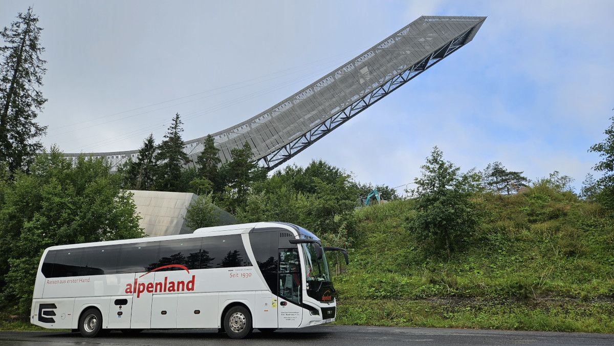 Der Alpenland Bus steht vor dem Holmenkollen, umgeben von Bäumen und Grün.