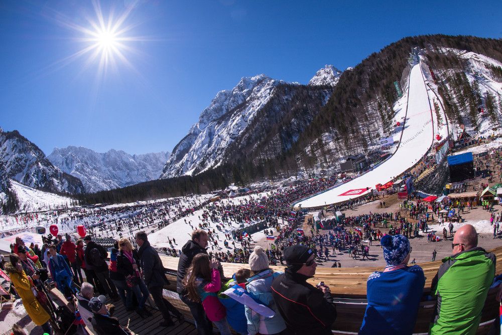 Ein sonniger Tag in den Alpen, zahlreiche Besucher beim Skispringen, umgeben von schneebedeckten Bergen.