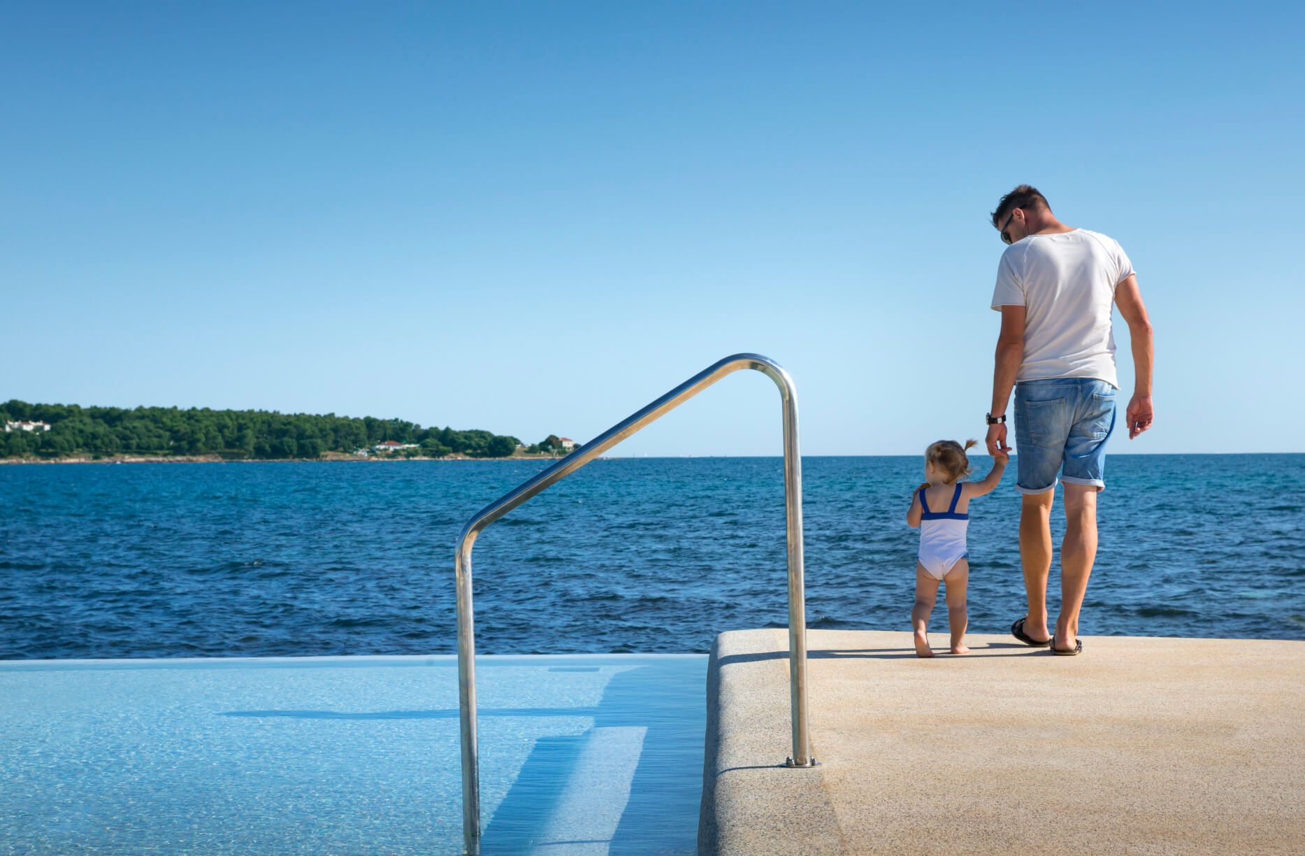 Ein Vater und sein Kleinkind stehen am Rand eines Pools, blicken aufs Meer und genießen den schönen Tag.