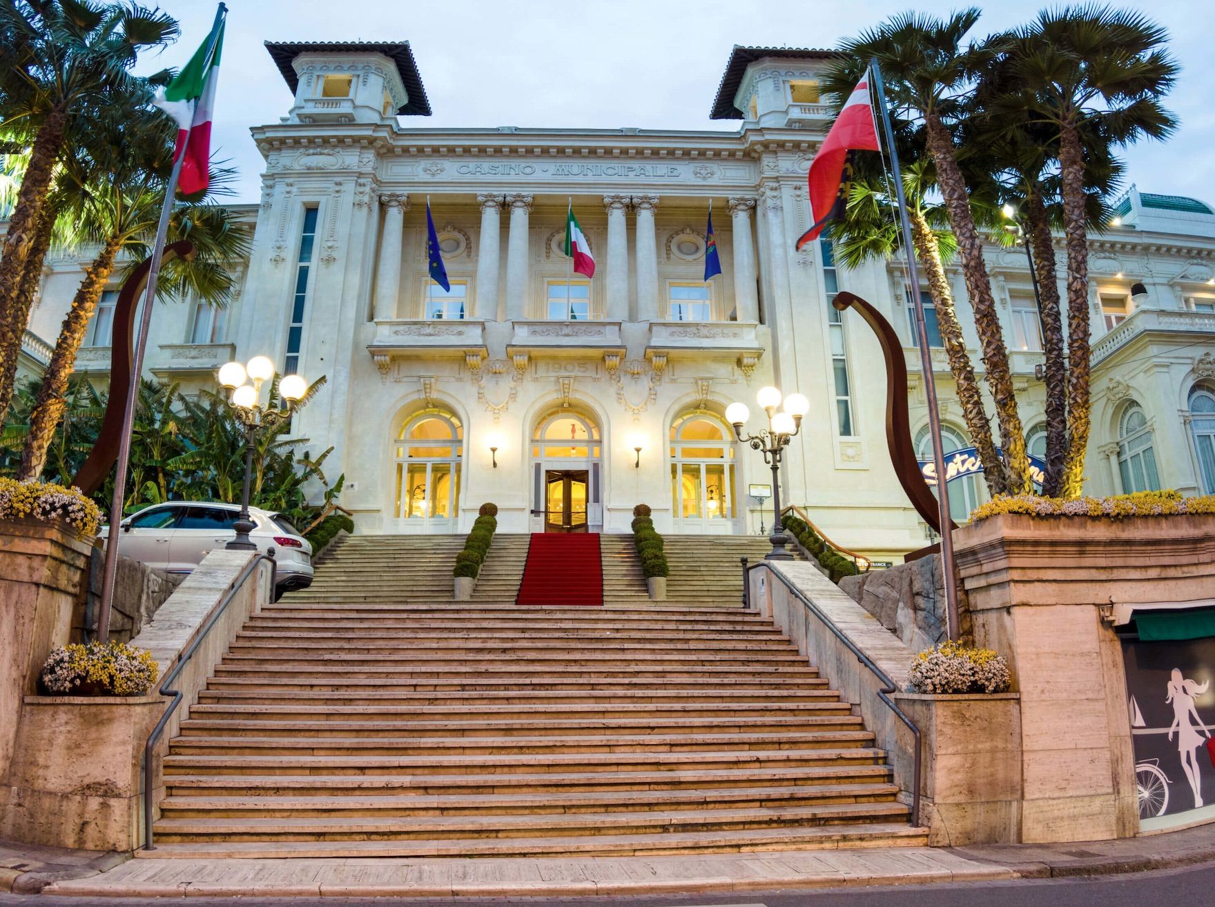 Fassade eines eleganten Casinos mit Treppen, Palmen und Fahnen, in sanften Farben bei Dämmerung beleuchtet.