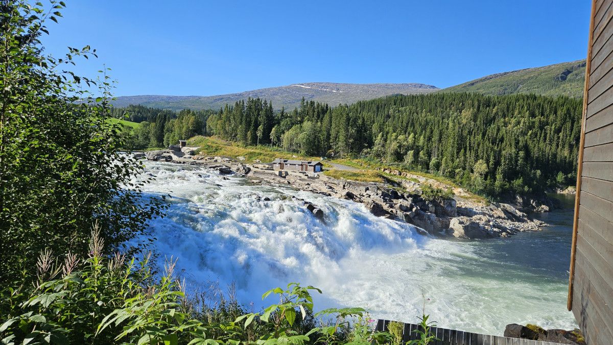 Ein malerischer Wasserfall umgeben von üppigem Grün und Bergen unter einem strahlend blauen Himmel.