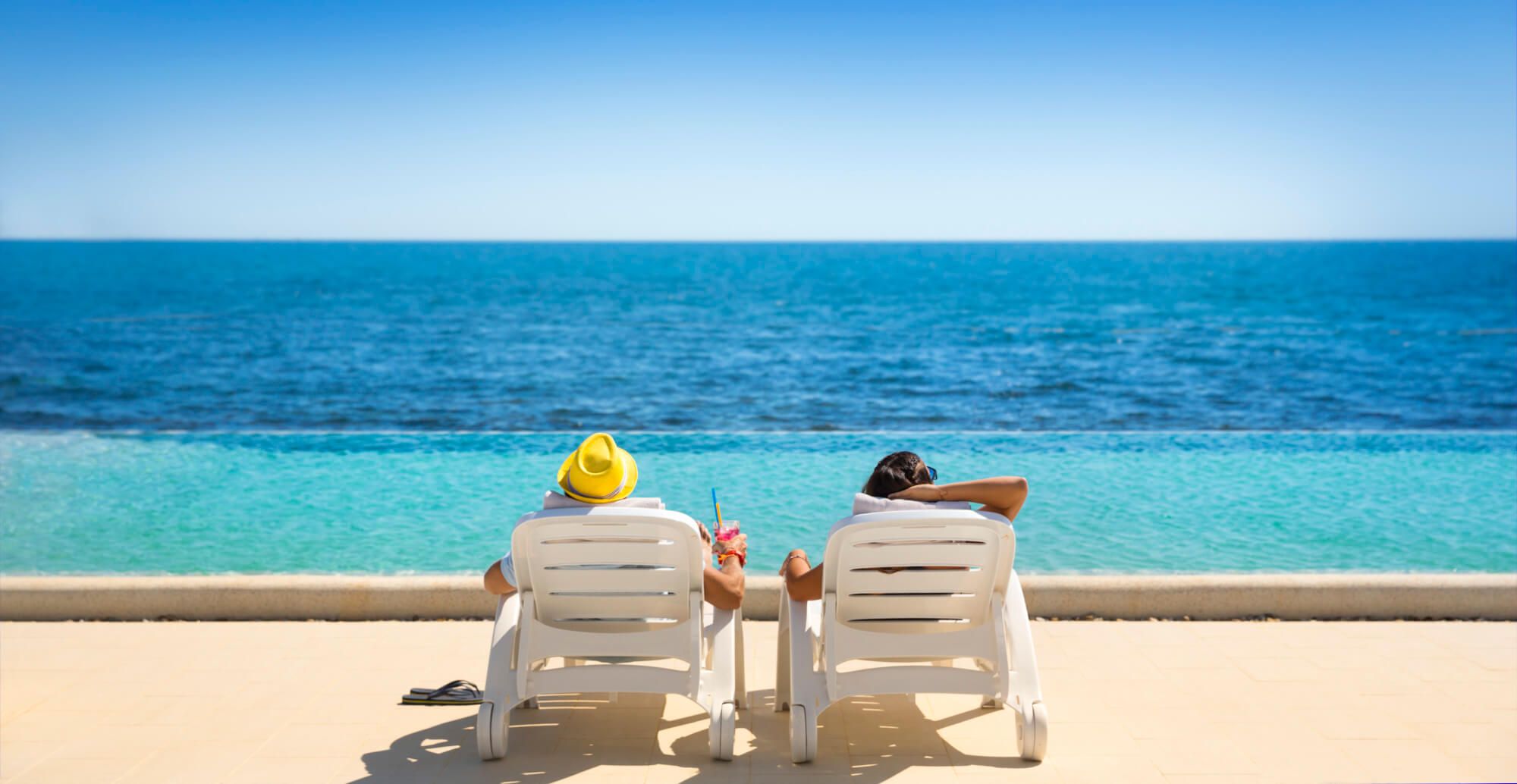 Ein Paar entspannt auf Liegen am Strand mit Blick auf das ruhige, azurefarbene Wasser und sonnigen Himmel.