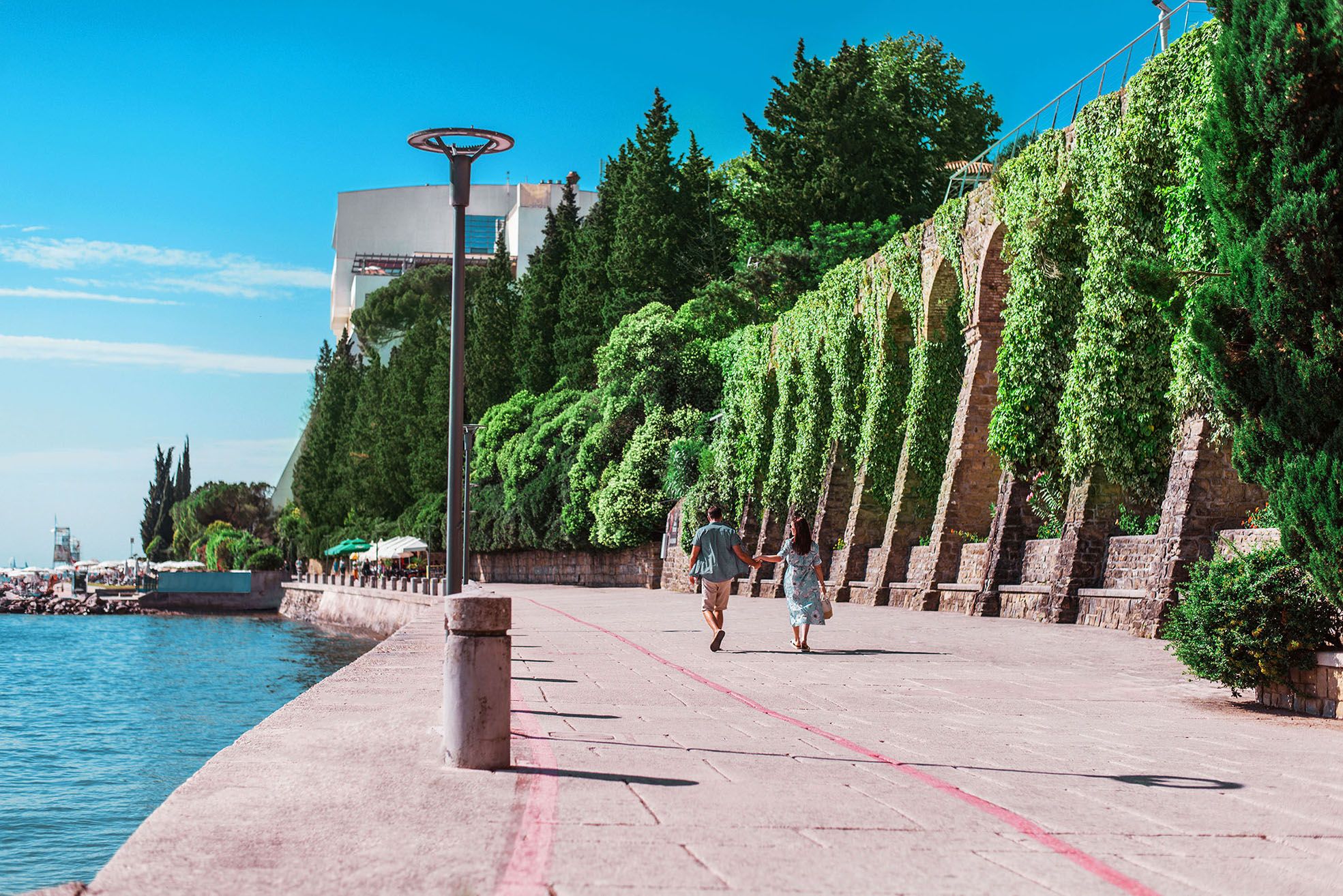 Ein Paar spaziert an einer sonnigen Uferpromenade, umgeben von üppigem Grün und einem blauen Himmel.
