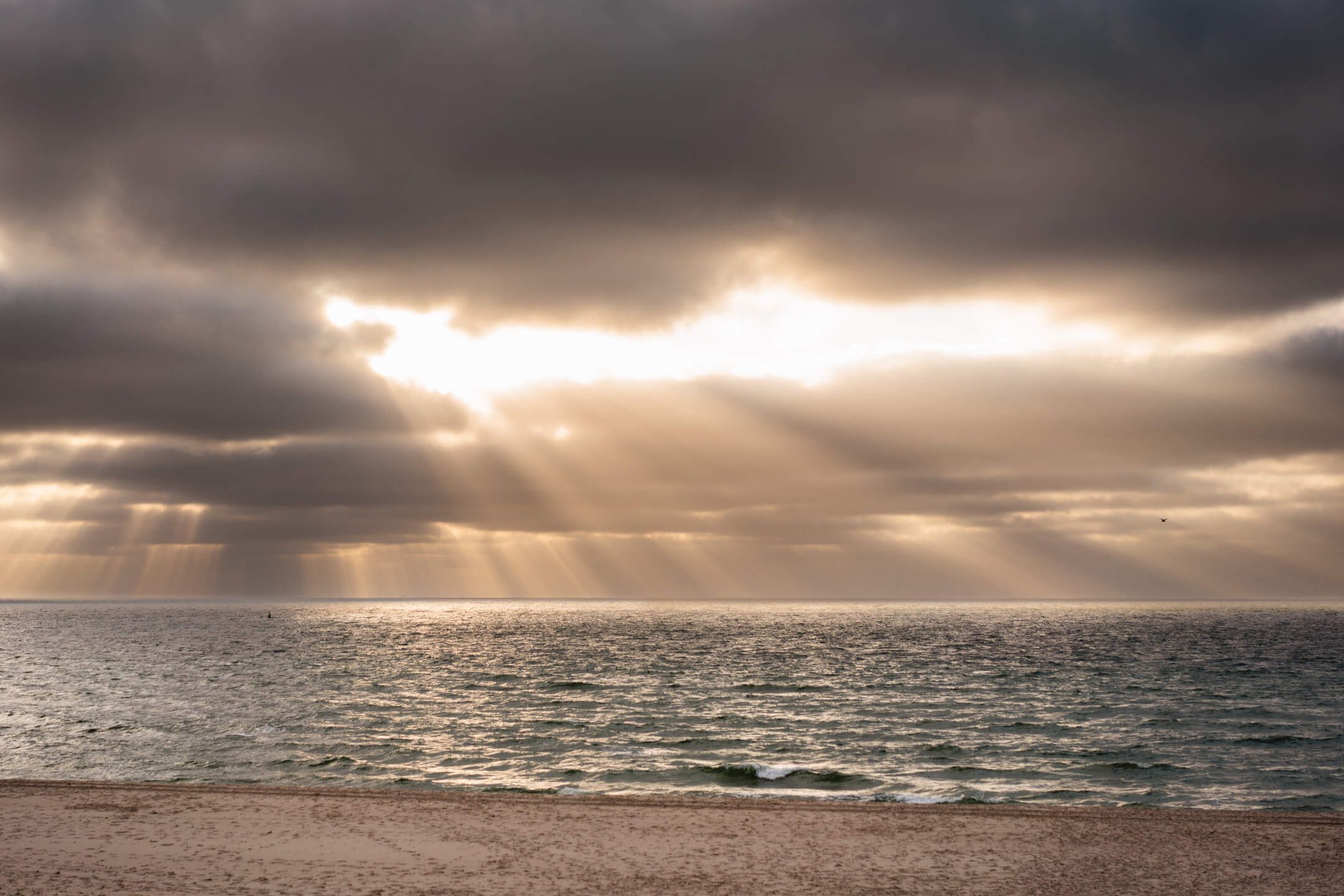 Dramatischer Himmel über dem Meer, mit Sonnenstrahlen, die durch die Wolken scheinen, und sanften Wellen am Strand.