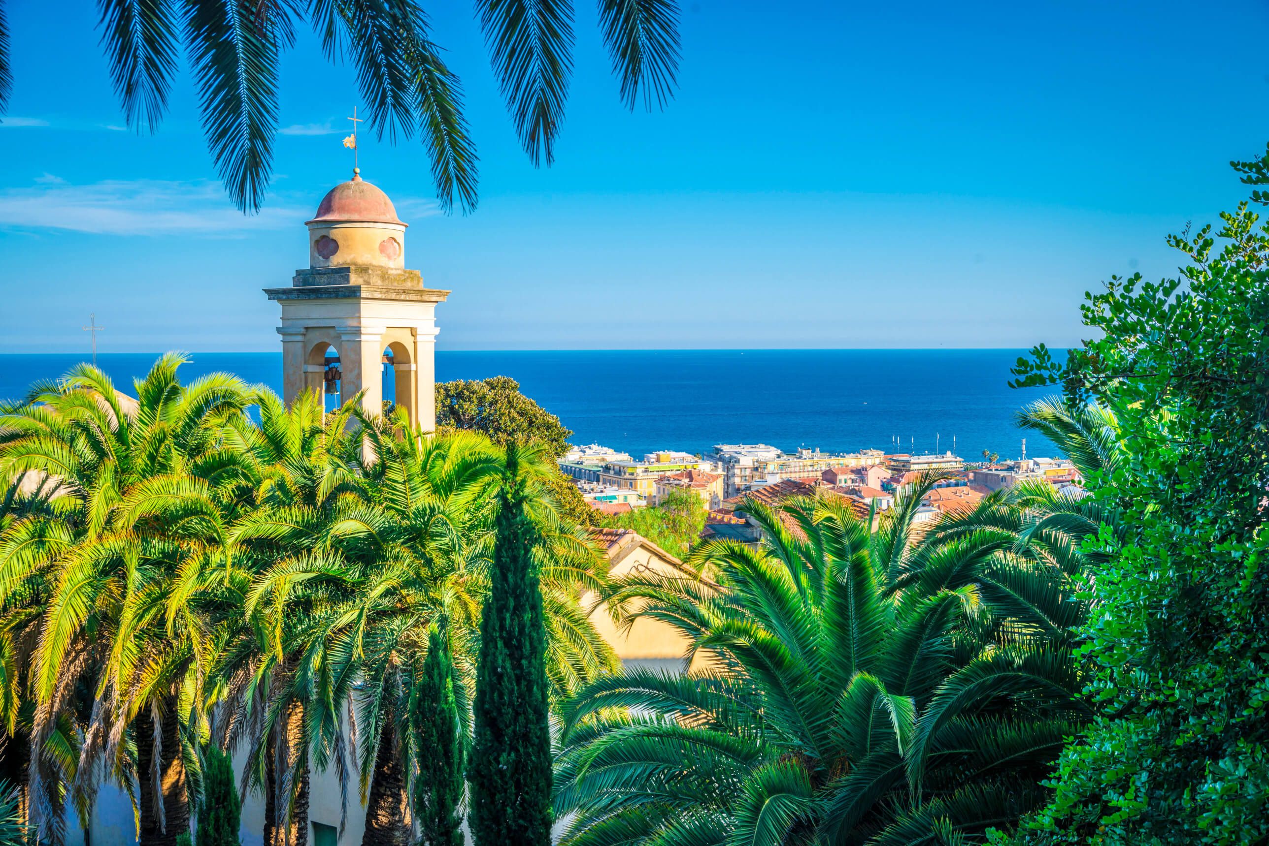 Blick auf einen Küstenort mit Palmen und einem Glockenturm vor blauem Himmel und dem Meer im Hintergrund.