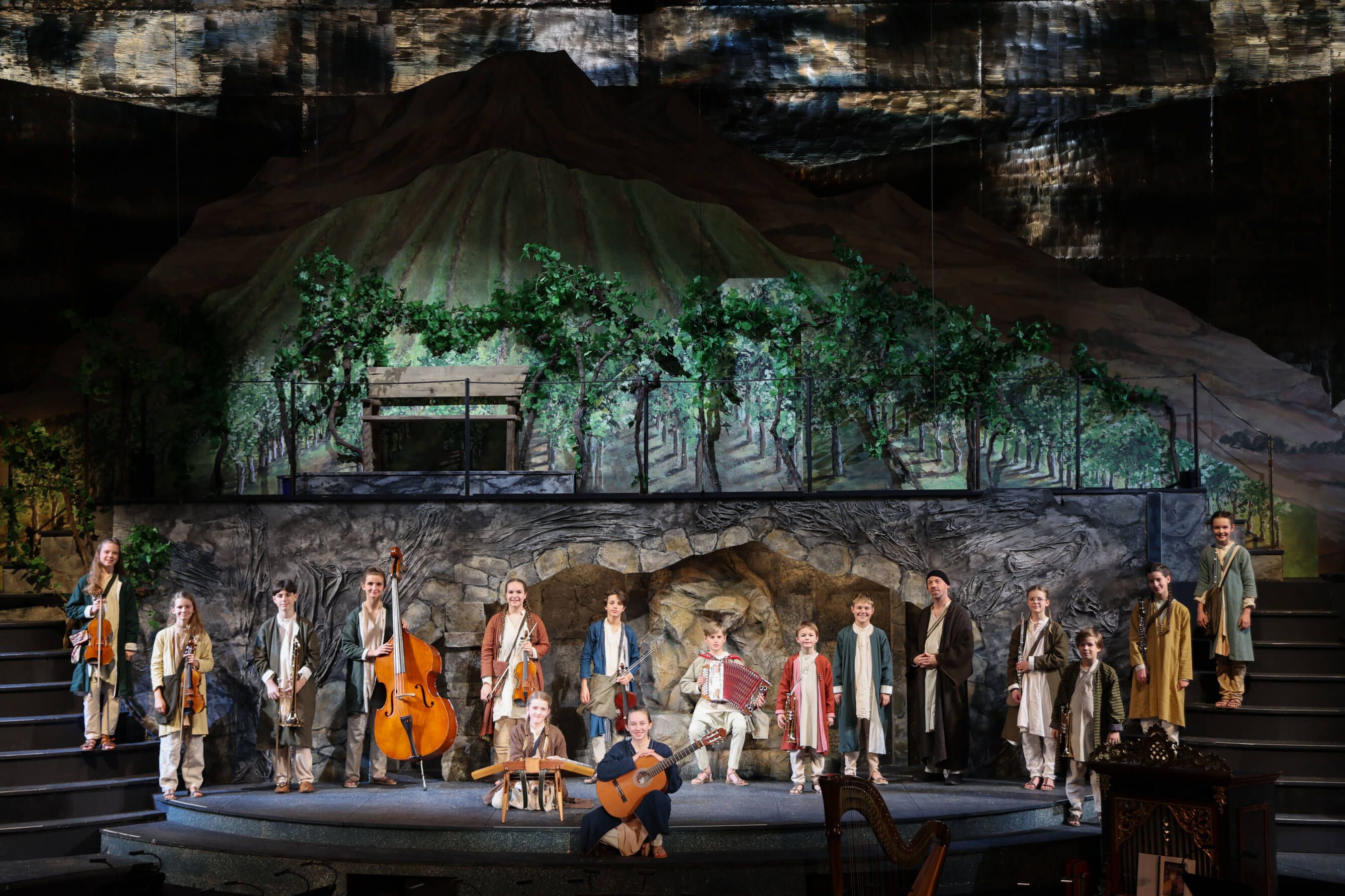 Die Bühne zeigt eine Gruppe von Kindern in historischen Kostümen mit Musikinstrumenten vor einer Berglandschaft.