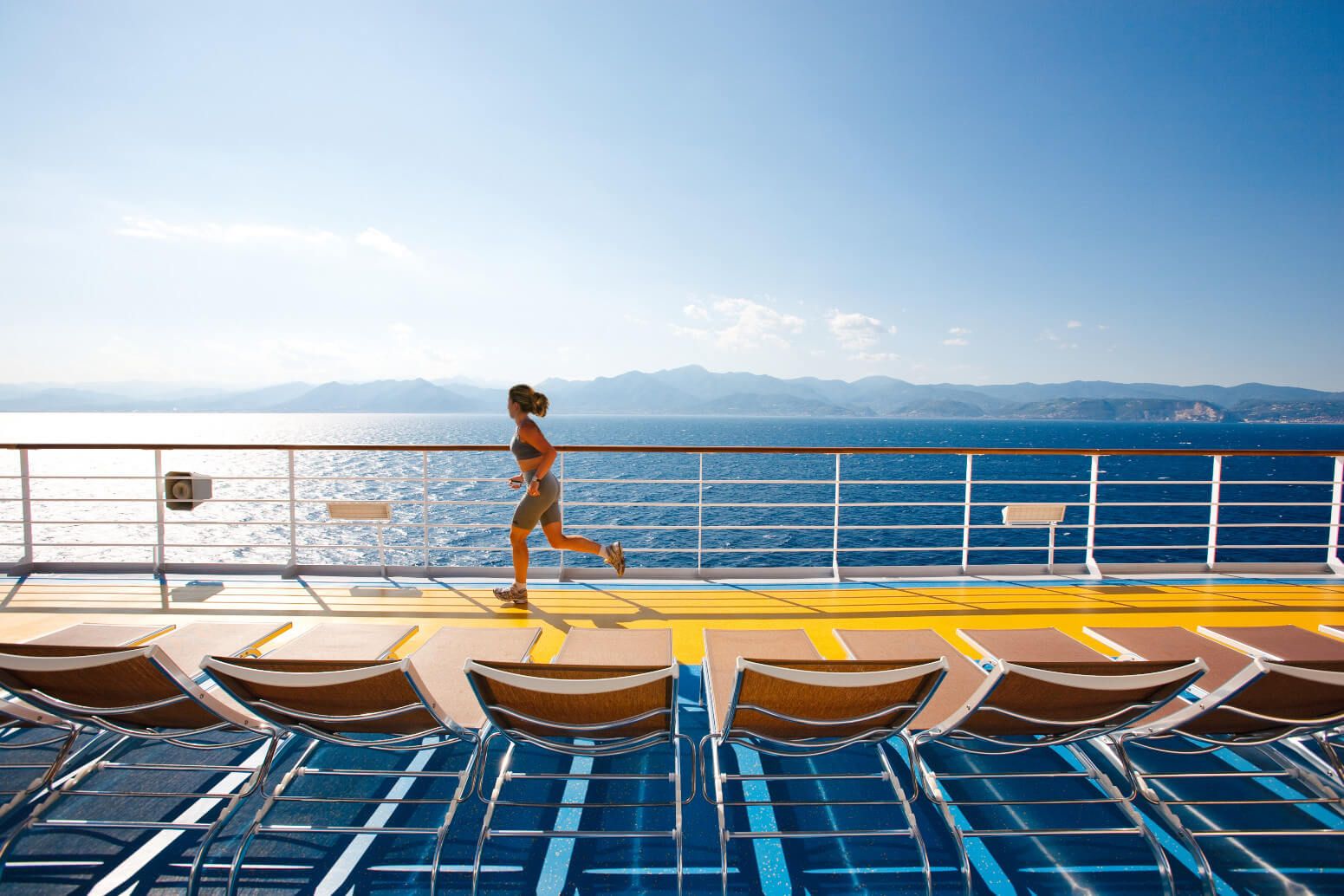 Eine Frau joggt auf einem Schiffdecks, mit Blick auf das Meer und die Berge im Hintergrund. Sonne scheint hell.