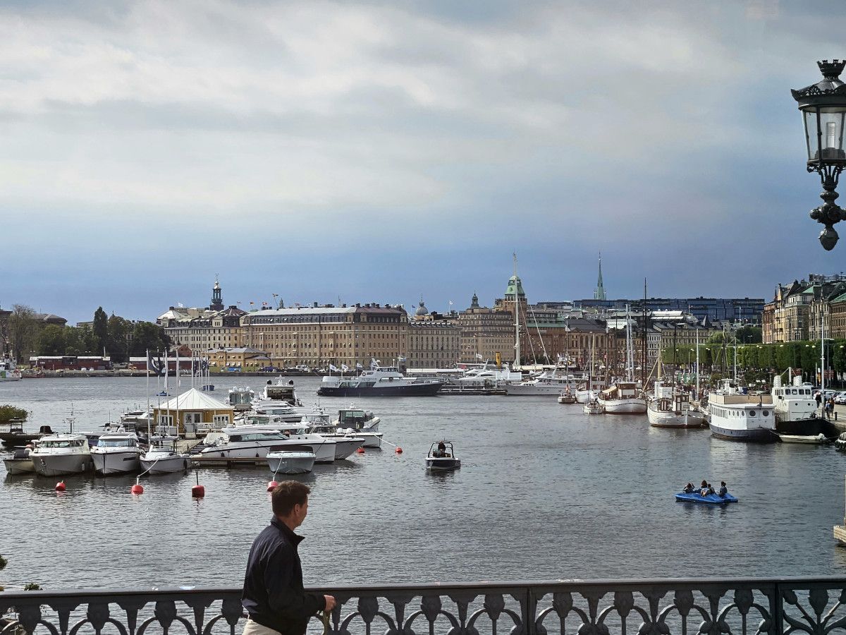 Ein Mann spaziert vorbei, während Boote im ruhigen Wasser einer Stadtlandschaft vor einem bewölkten Himmel liegen.