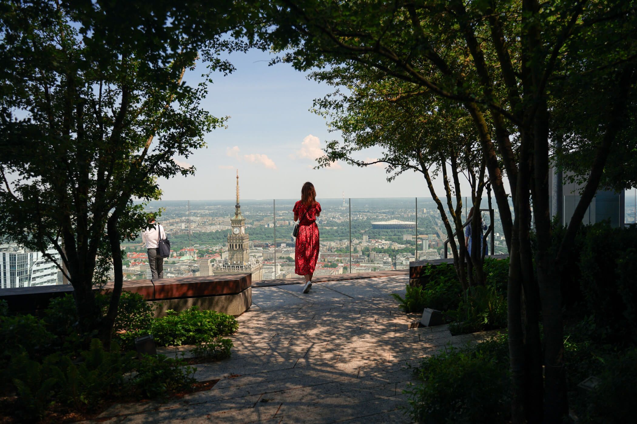 Eine Frau in einem roten Kleid steht an einer Aussichtsterrasse mit Blick auf die Stadt und den markanten Turm.