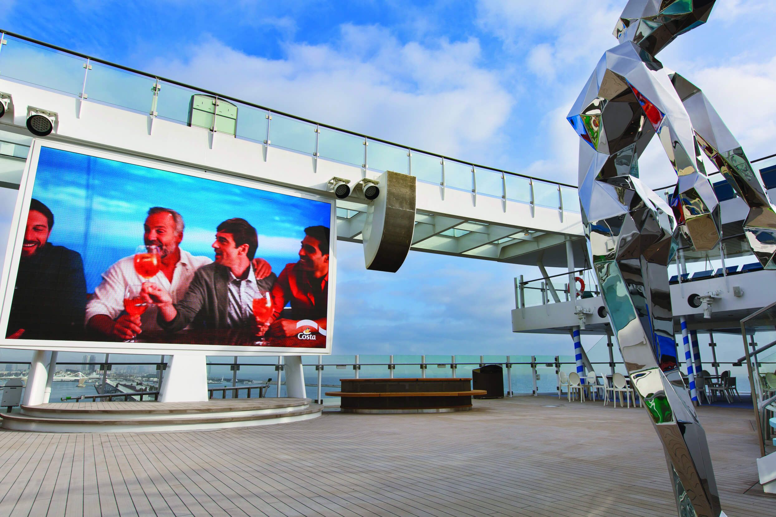 Ein moderner Außenbereich mit großem Bildschirm und einer kunstvollen Skulptur auf einem Schiff bei klarem Himmel.