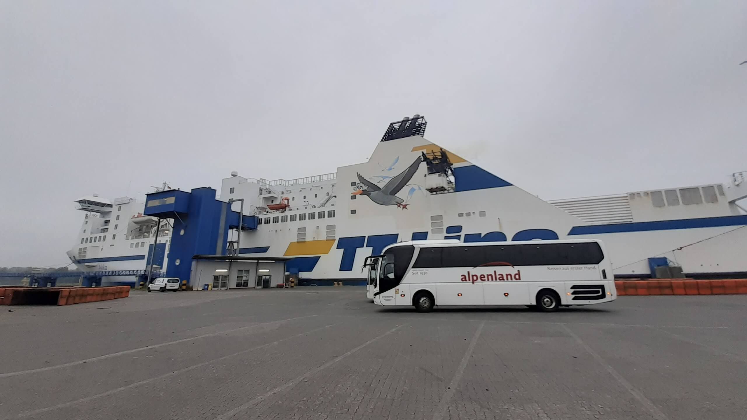 Ein großes Fährschiff mit buntem Design steht am Hafen, ein Bus ist im Vordergrund sichtbar. Grauer Himmel.
