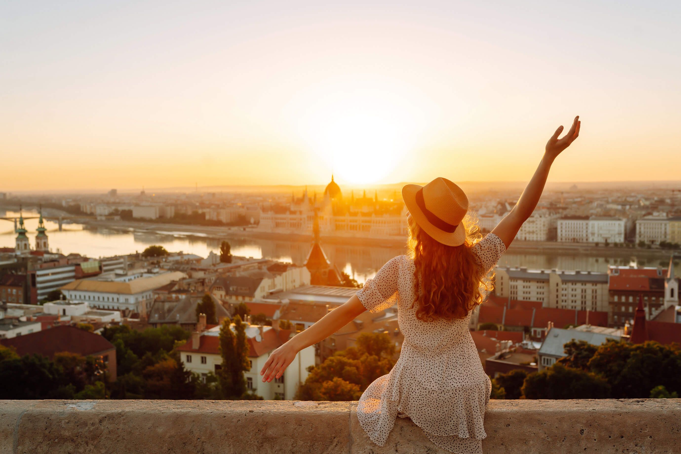 Eine Frau mit Hut sitzt am Rand, während die Sonne über der Stadt aufgeht. Eine malerische Aussicht!