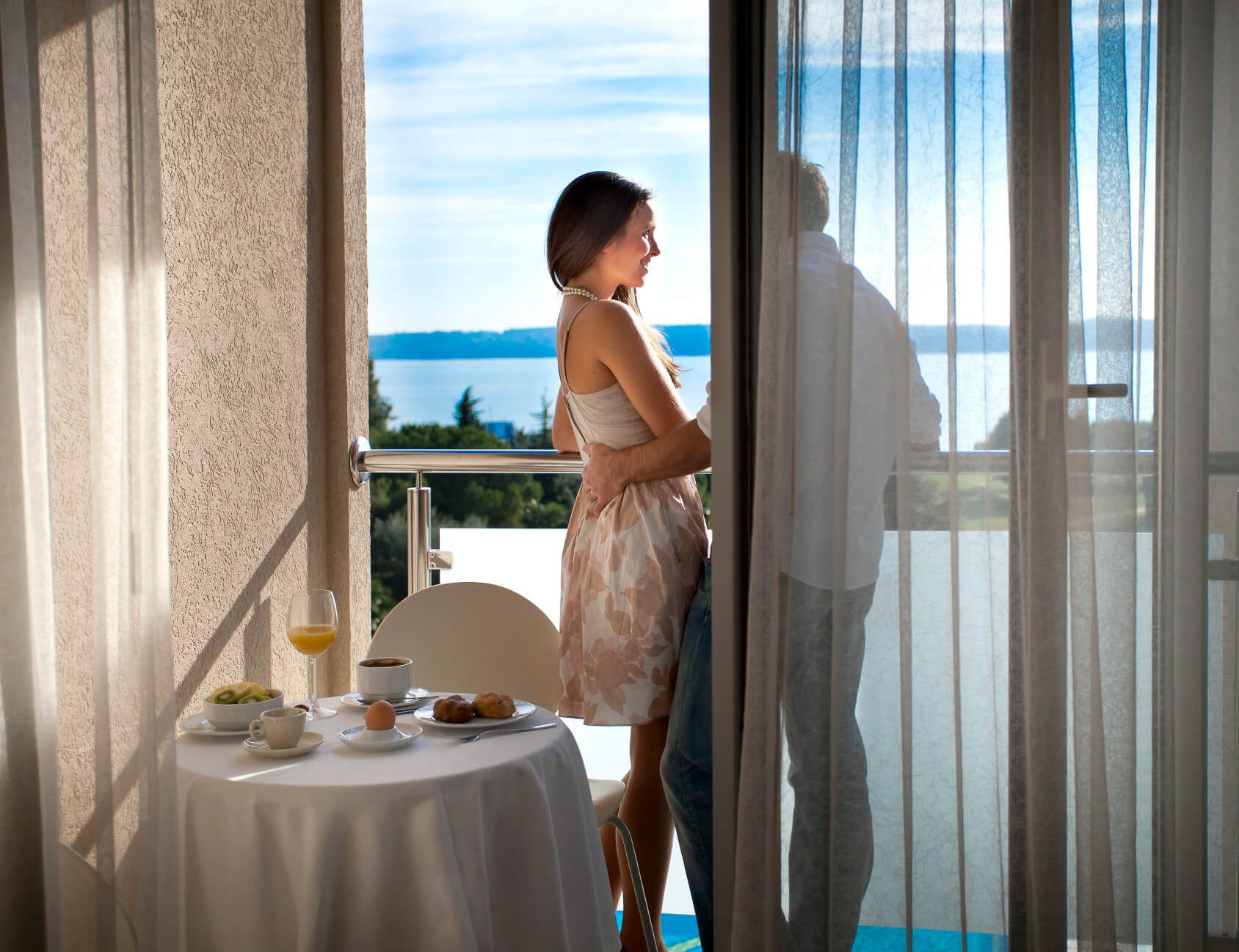 Ein Paar steht auf dem Balkon, genießt den Ausblick auf das Wasser und ein Frühstückstisch ist vorbereitet.