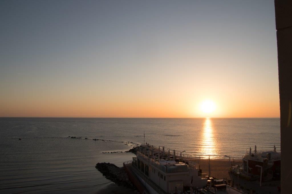 Sonnenuntergang über dem Meer, sanfte Wellen, ein Boot am Ufer, warme Farben am Himmel.