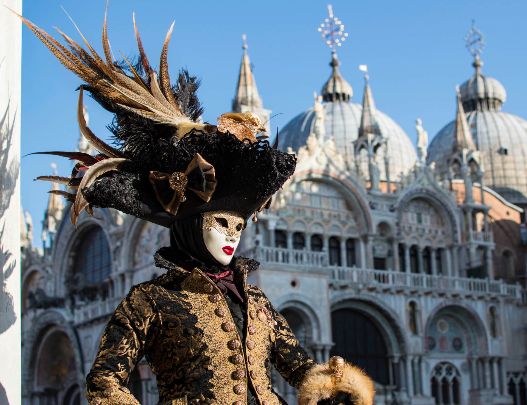 Eine elegante Person in einer aufwendigen Maske und Kostüm steht vor einer beeindruckenden historischen Kulisse in Venedig.
