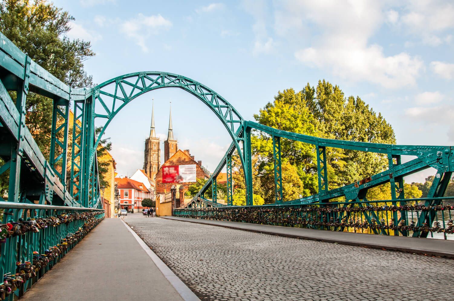 Eine malerische Brücke mit Liebe-Schlössern, im Hintergrund zwei beeindruckende Kirchtürme und herbstliche Bäume.