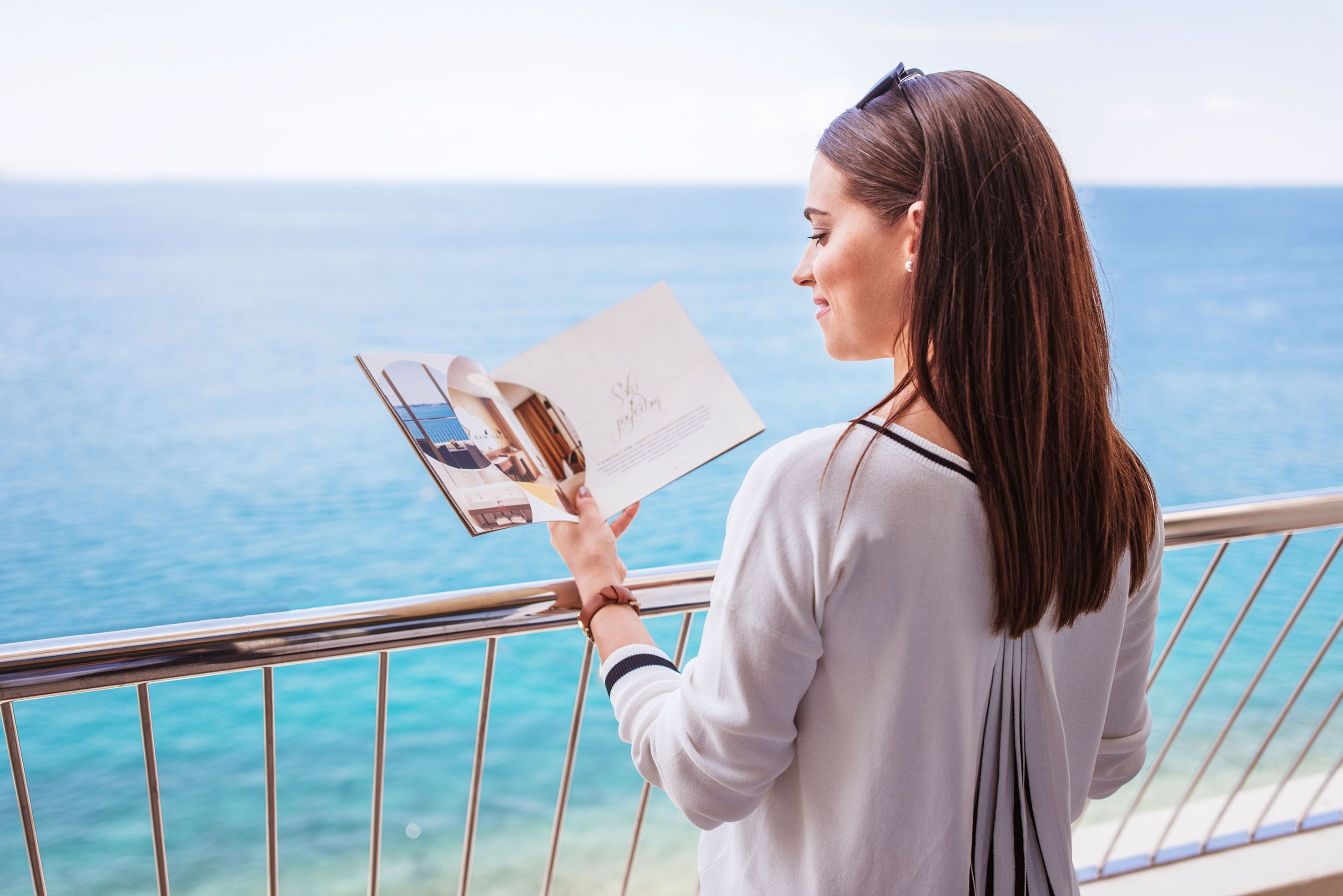 Eine Frau liest entspannt eine Broschüre auf einem Balkon mit Meerblick.