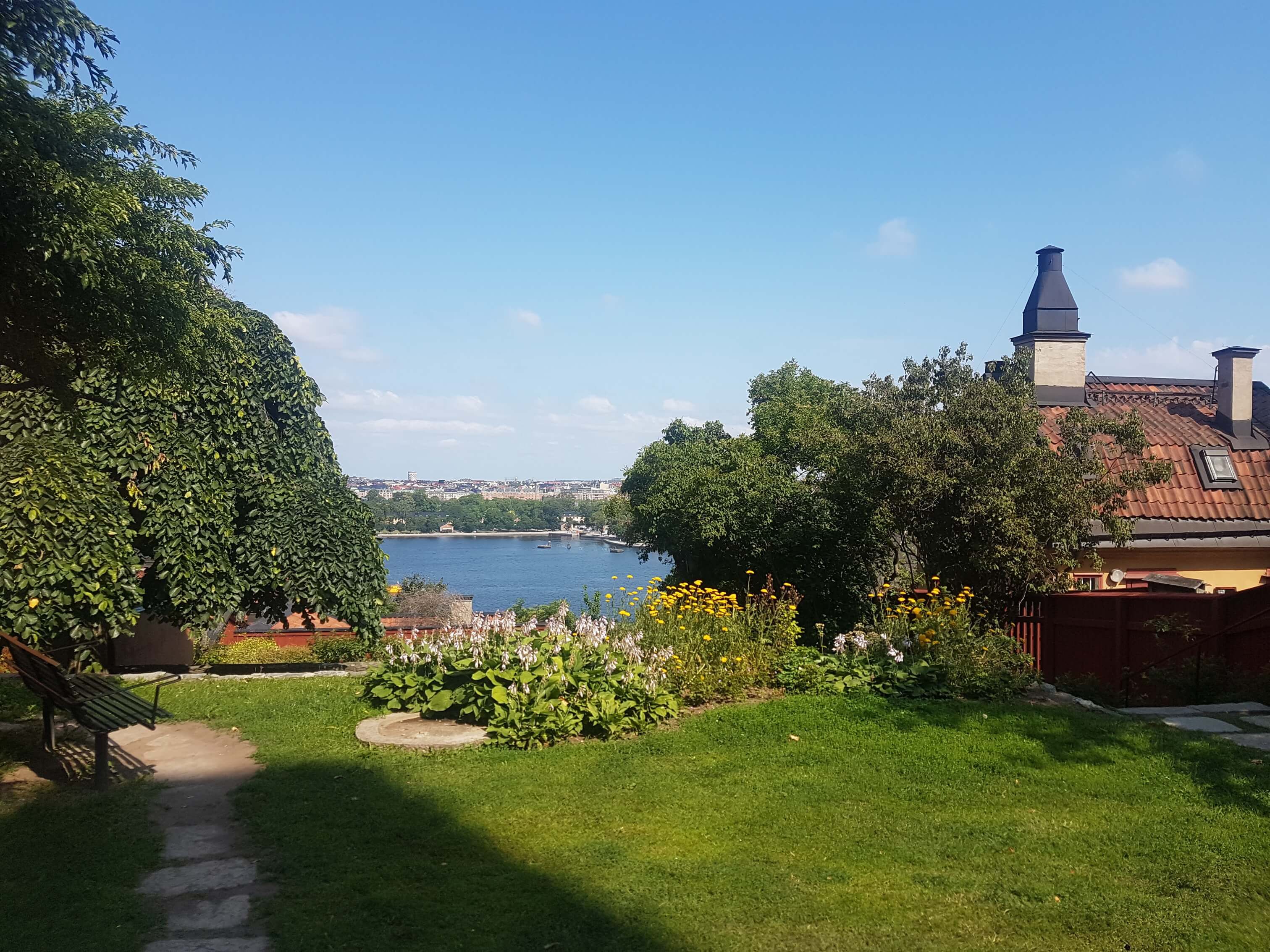 Ein schöner Garten mit bunten Blumen, Blick auf das Wasser und Häuser im Hintergrund unter blauem Himmel.