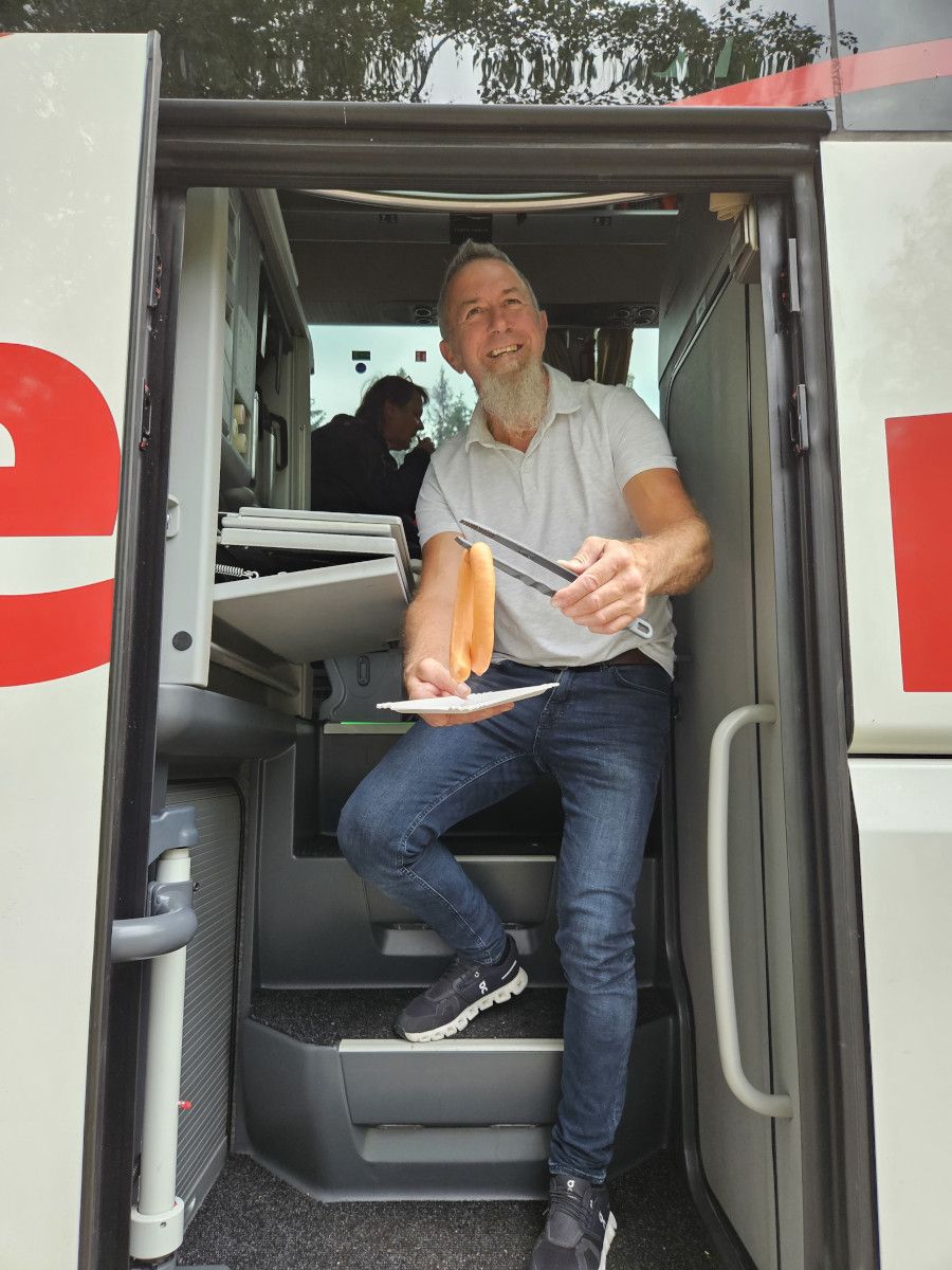 Ein Mann sitzt in einem Fahrzeug und hält eine Wurst mit einer Gabel, während er lächelt.