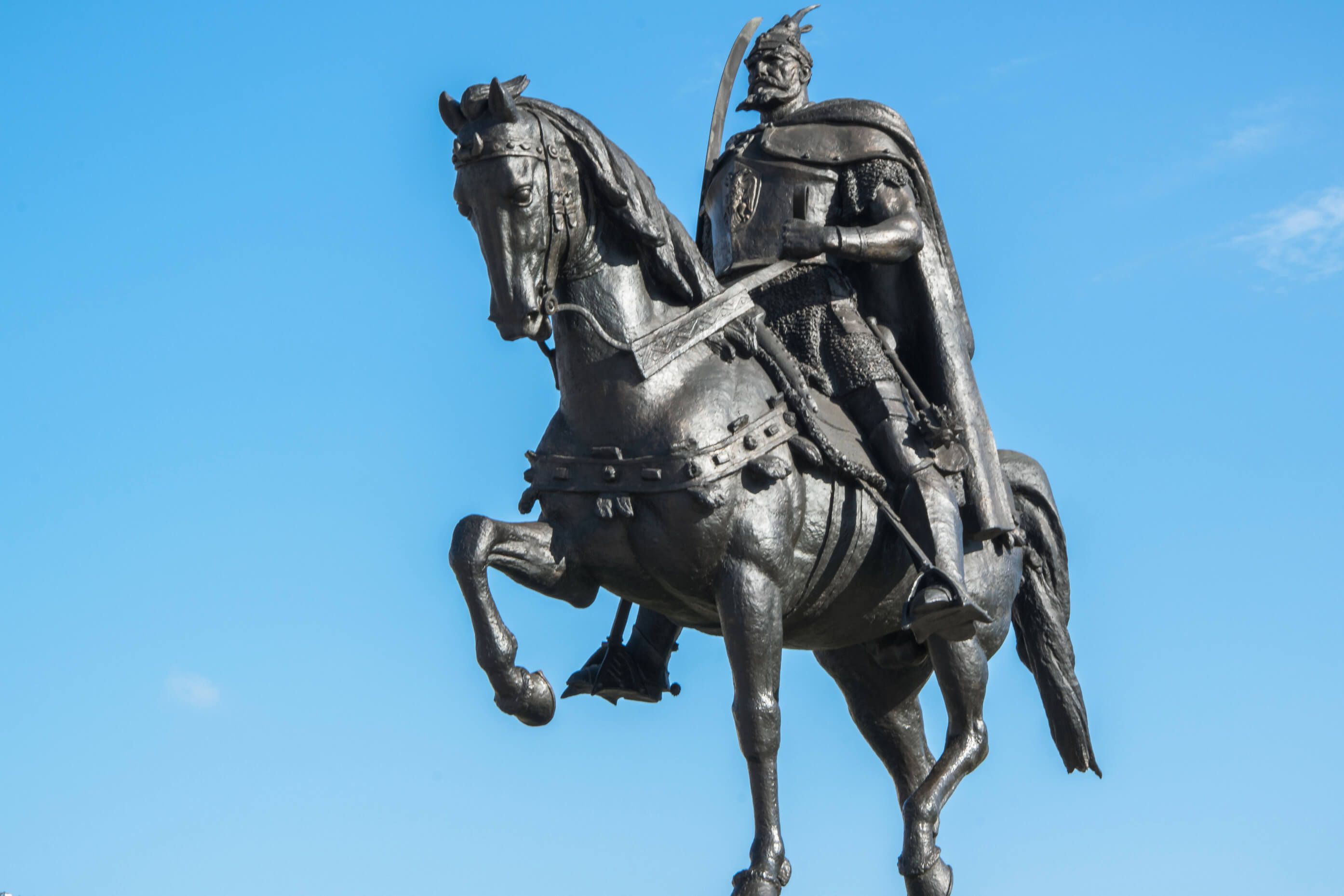 Eine bronze Statue eines kriegerischen Mannes auf einem Pferd vor blauem Himmel.