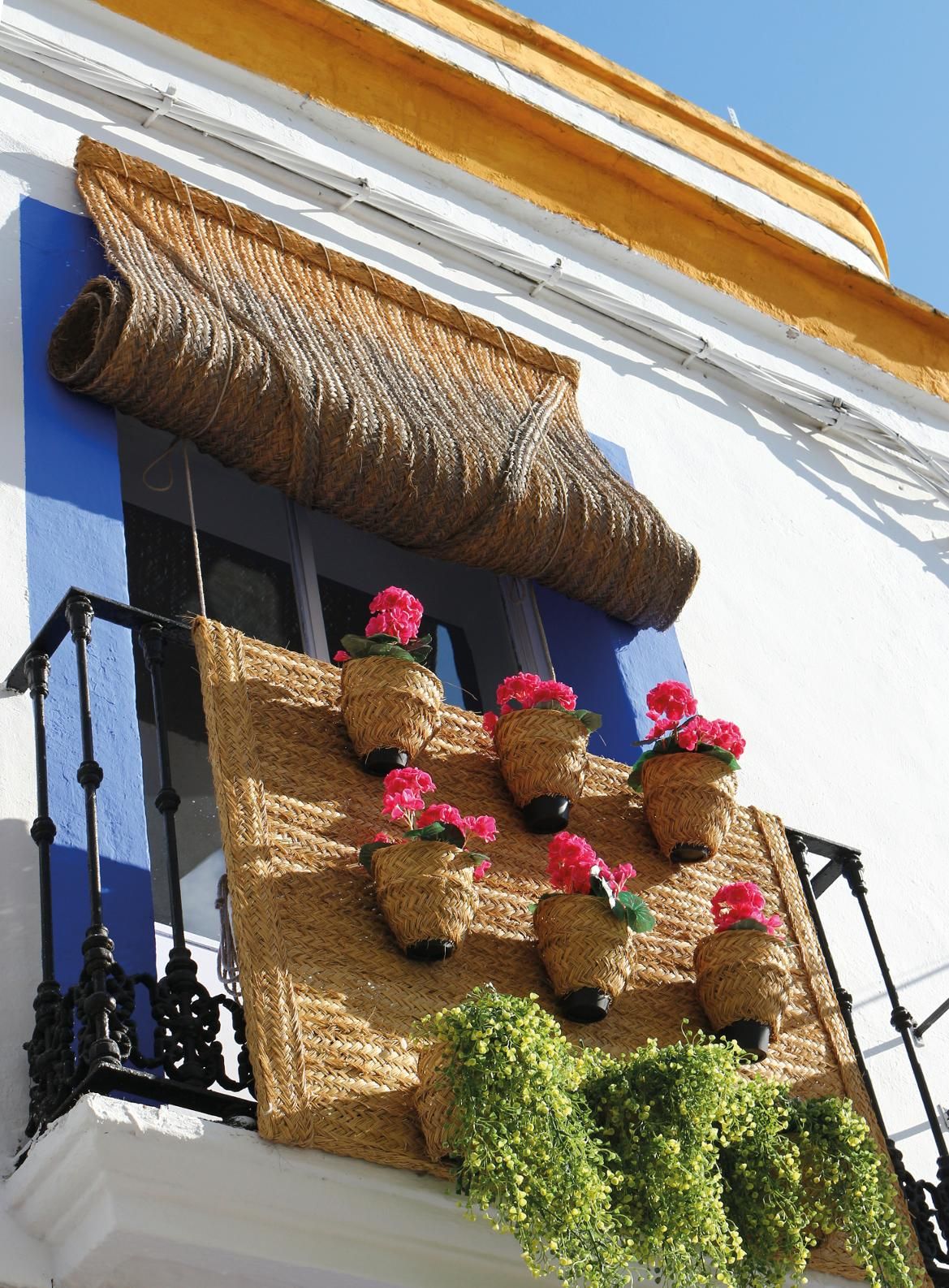 Ein Balkon mit blauen Fensterrahmen, geflochtenen Töpfen und bunten Blumen in einem sonnigen Ambiente.