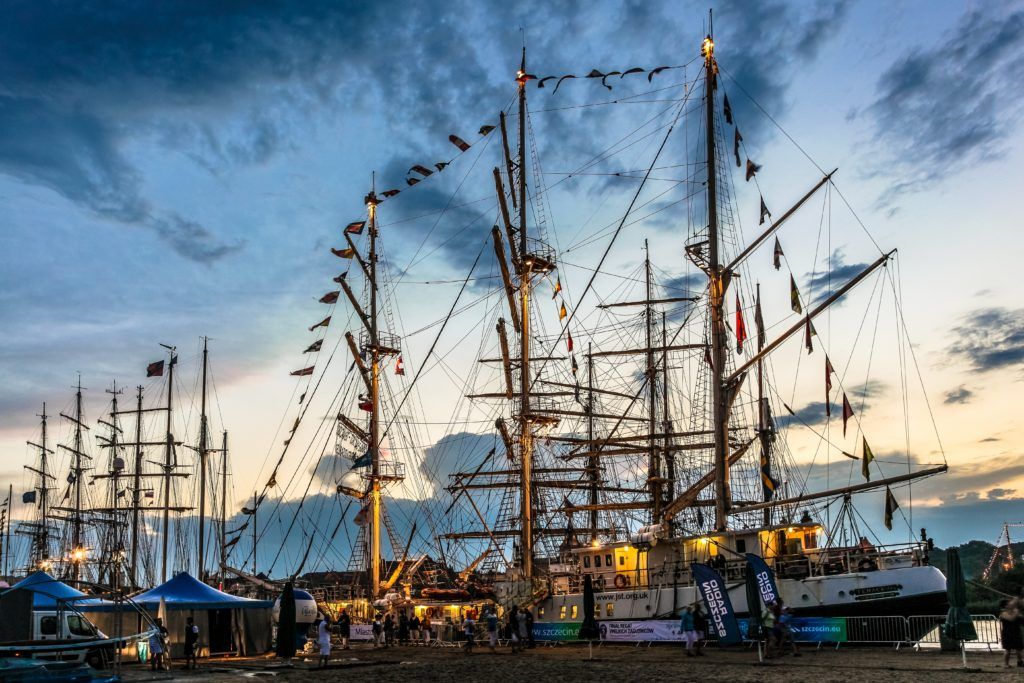 Eine Szene mit historischen Segelschiffen im Hafen unter bewölktem Himmel bei Dämmerung, Bunte Flaggen wehen.